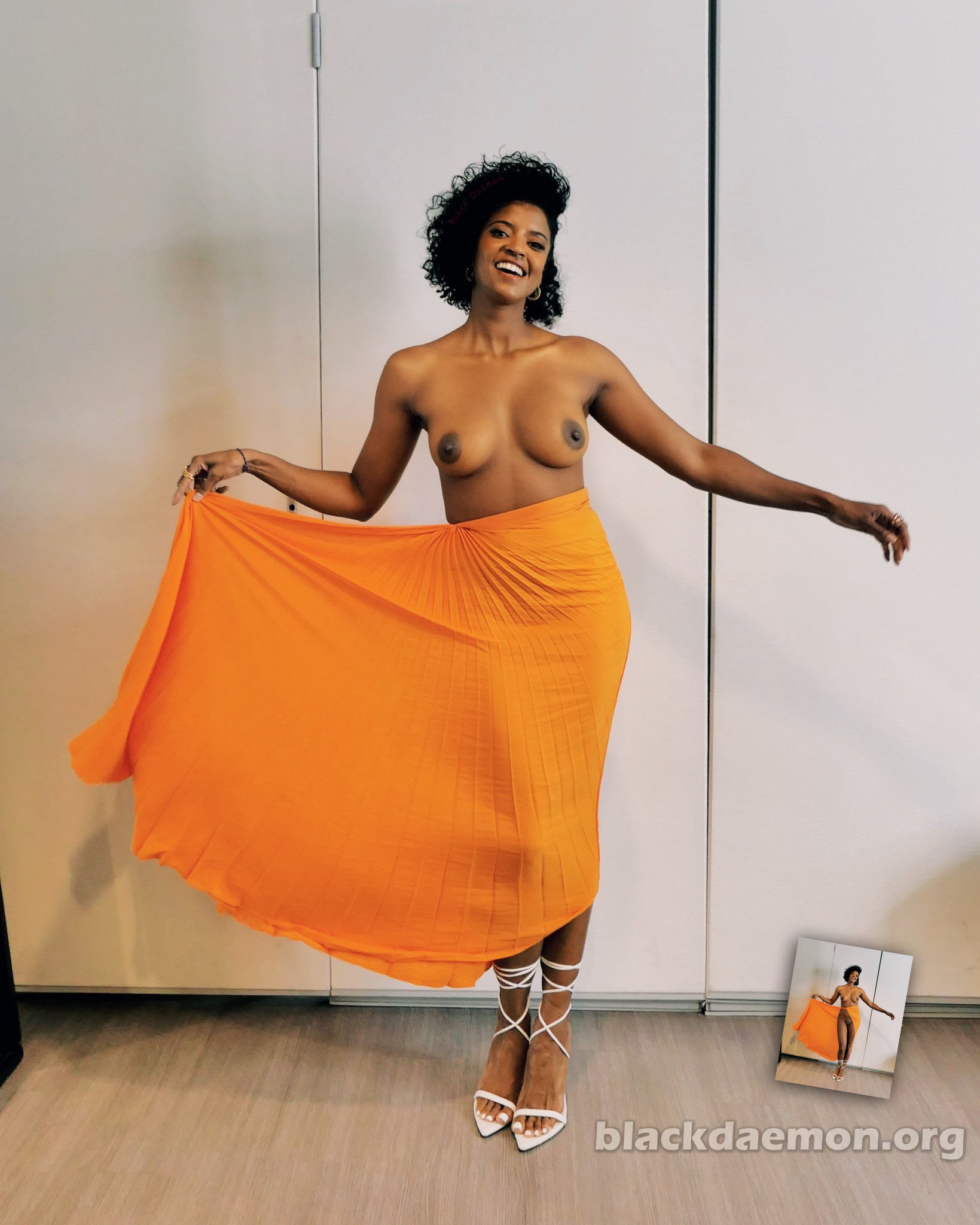 Renée Elise Goldsberry nude in an orange skirt is standing in front of a white wall.